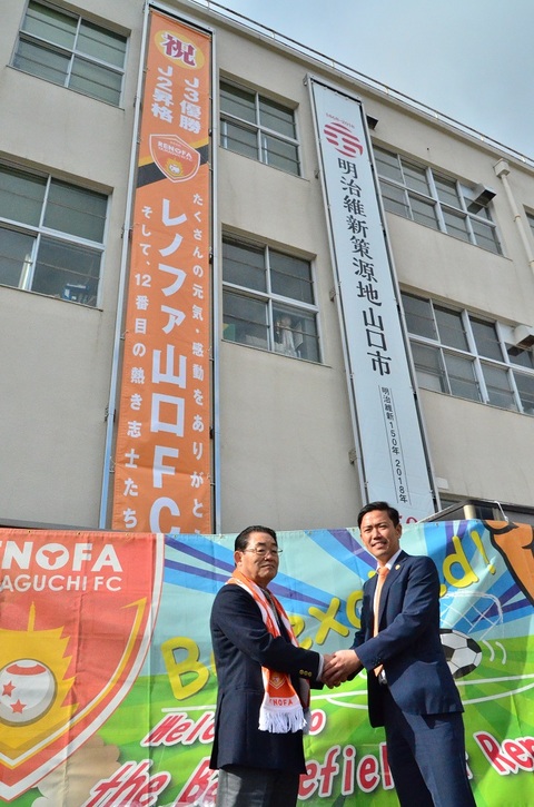 懸垂幕の前で握手する吉田副市長(左)と石原ゼネラルマネジャー(右)