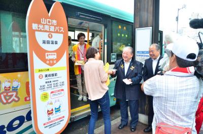 第1便の乗客に記念品を渡す市長、松村防長交通社長と車内でお出迎えする観光ボランティア