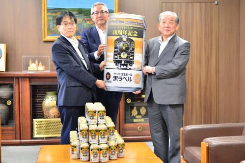 記念ラベル缶のパネルを手にする市長と忠沢中四国本部長（中央）、真辺部長（左）